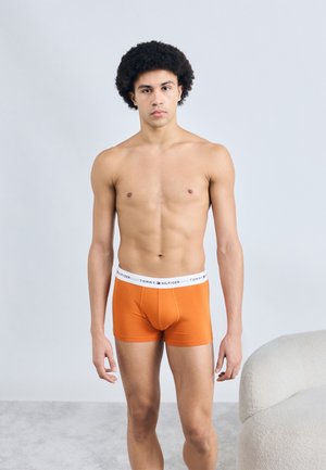 Oranje katoenen boxershorts met een witte tailleband met logo. Het model staat tegen een lichtgrijze achtergrond en toont een aansluitend ontwerp.