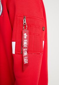 Felpa rossa con una tasca a zip sulla manica, dotata di un'etichetta rossa con testo bianco "RIMUOVERE PRIMA DEL VOLO". Texture liscia in cotone.