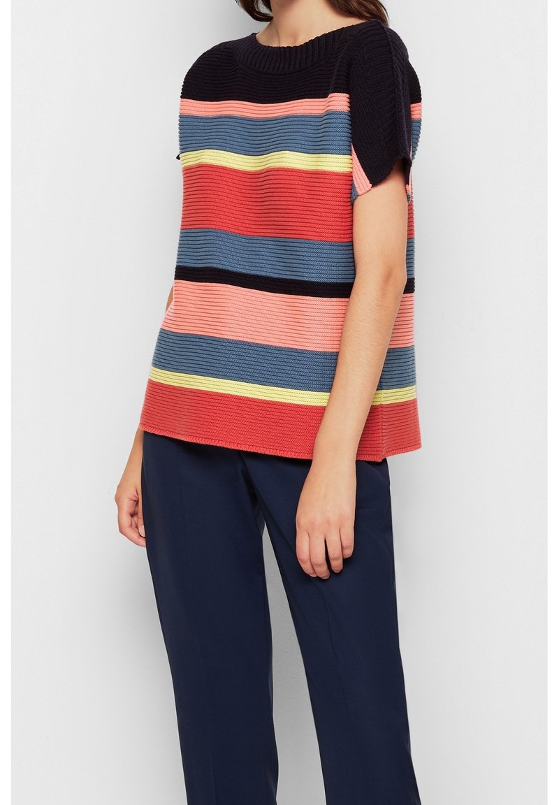 Vrouw in een kortemouwen gebreide top met horizontale strepen in zwart, roze, blauw, geel en rood, gecombineerd met donkerblauwe broeken.