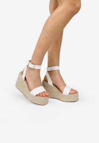 Sandalias de cuña blancas con suelas de yute trenzado estilo espadrille. Presentan correas anchas, hebilla en el tobillo y un diseño de punta abierta. Textura suave.