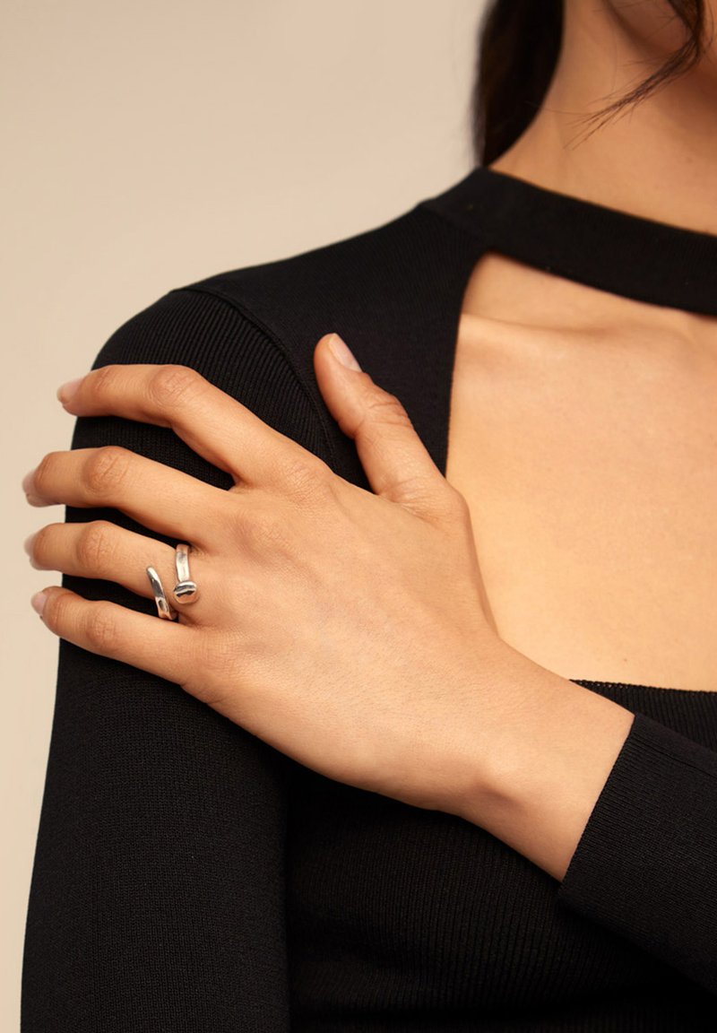 Anillo de plata con un diseño abstracto, que comprende elementos suaves y curvados. Top de manga larga negro con textura acanalada, que muestra un detalle de corte en el hombro.