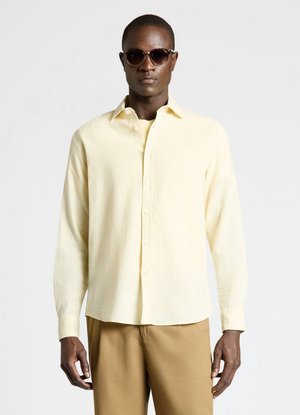 Man wearing dark sunglasses, light yellow long-sleeve button-up shirt, and tan pants standing against plain light background.