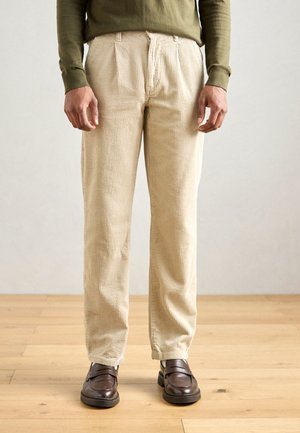 Pantalons en velours côtelé beige avec une texture à côtes verticales, featuring des plis à l'avant et une coupe droite. Portés avec des chaussures brunes à enfiler.