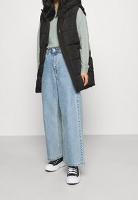 Svart vadderad väst, ljusgrön tröja och vida blå jeans. Skor inkluderar svartvita sneakers med tjock sula och skosnören.