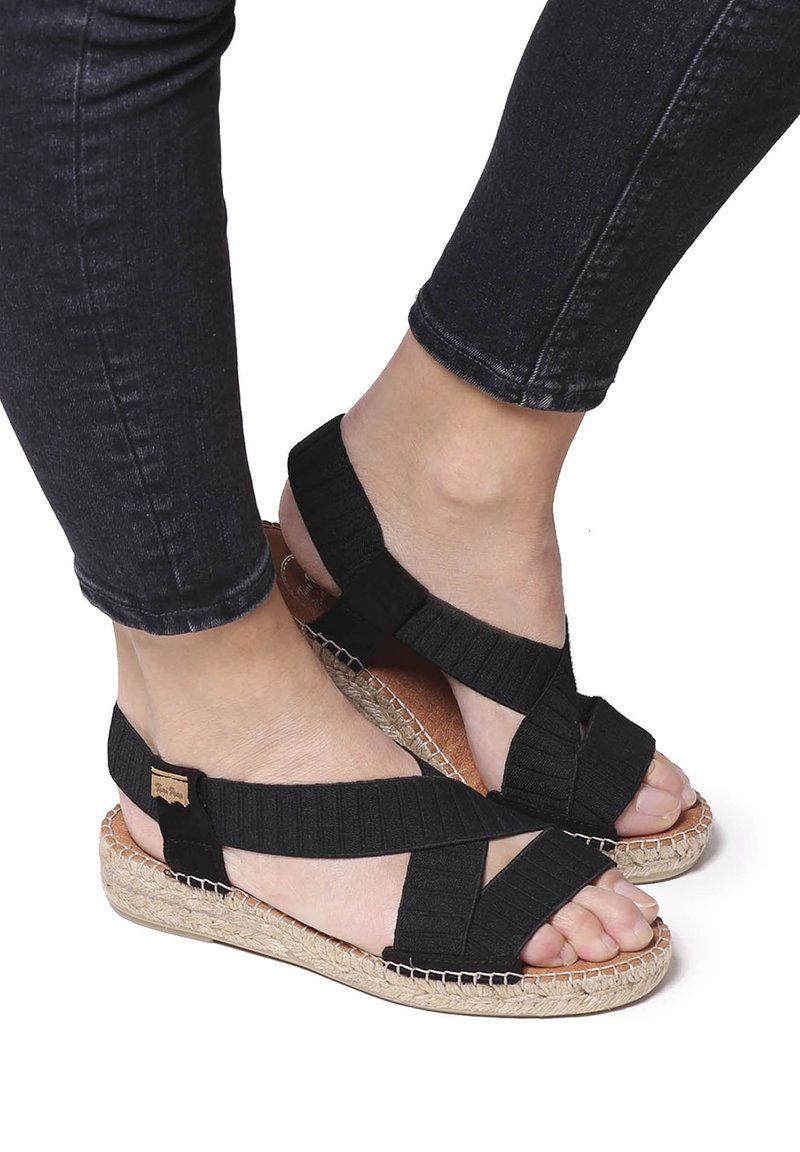 Piedi che indossano sandali neri con cinturini elasticizzati e suole in espadrilles intrecciate, abbinati a jeans skinny neri.