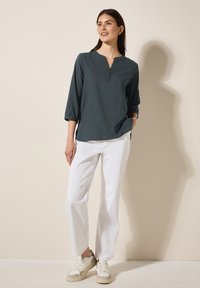 Dunkelbluse in Teal mit V-Ausschnitt und dreiviertel Ärmeln, ausgestattet mit einer Knopfleiste. Kombiniert mit weißen geraden Hosen und Sneakers.