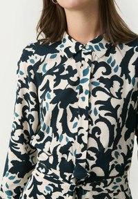 Blusa a fantasia con un design floreale blu navy e crema, con colletto button-down e maniche lunghe. Il tessuto appare leggero e morbido.