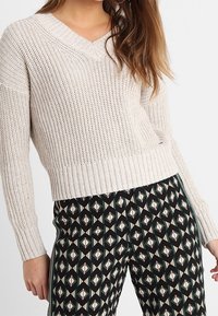 Mujer con un suéter de punto beige con escote en V y pantalones negros con un patrón geométrico de diamantes y franjas laterales.