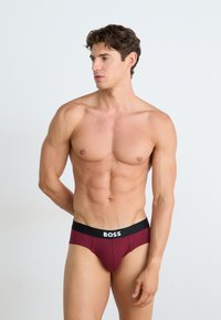 Caleçons maroon avec une ceinture noire arborant le logo "BOSS". Texture lisse, forme ajustée et design taille basse.