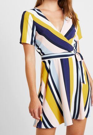 Kobieta ubrana w sukienkę kopertową z krótkim rękawem w ukośne paski w kolorach żółtym, granatowym, jasnoniebieskim, brzoskwiniowym i białym, przewiązaną w talii paskiem.
