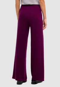Donna in pantaloni larghi viola e scarpe nere, vista da dietro su uno sfondo grigio chiaro uniforme.