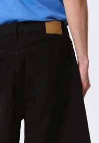 Personne portant un short en denim noir avec des poches arrière et un patch en cuir marron marqué "WEEKDAY", associé à une chemise bleue.
