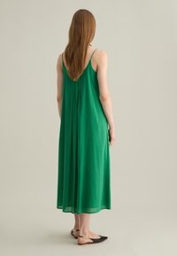 Abito maxi senza maniche verde con una silhouette fluida, pieghe sul retro e tessuto leggero; abbinato a ballerine nere con accenti dorati.