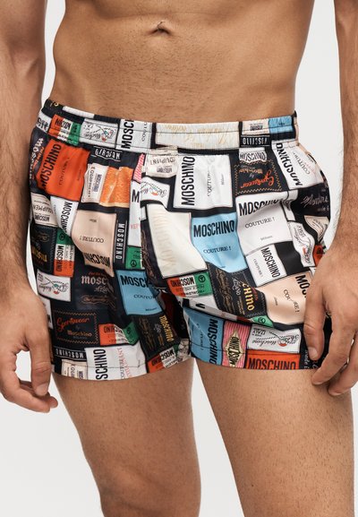 Άνδρας που φορά πολύχρωμα σορτς Moschino με πολλαπλά σχέδια ετικετών σε διάφορα χρώματα, στέκεται με τα χέρια χαλαρά δίπλα στο σώμα.