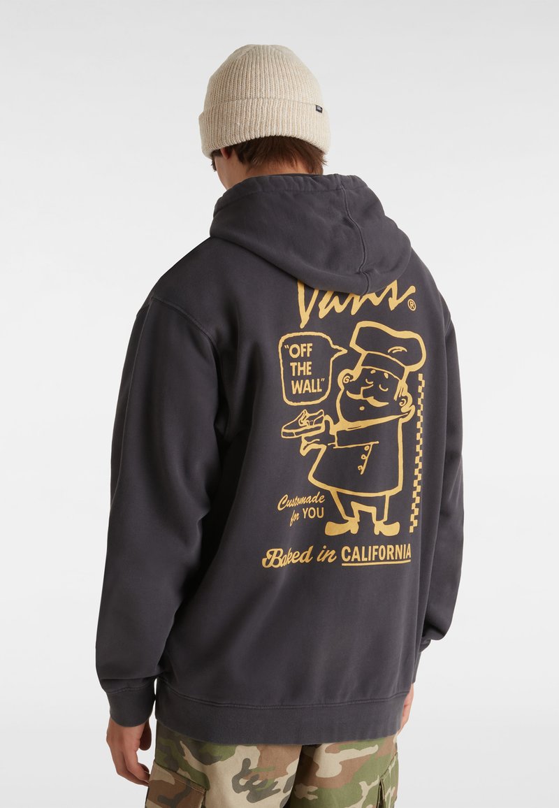 Felpa grigia scura con design grafico giallo sul retro, che presenta un personaggio chef e del testo. Polsini e orlo a costine. Indossata con pantaloni mimetici.