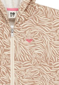 Beige fleecová mikina na zips s ružovým kvetinovým a zebrovým vzorom. Obsahuje malú ružovú srdcovú nálepku a kontrastný zips.