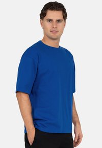 T-shirt bleu en coton à col rond, manches courtes, coupe décontractée et coutures plates. Porté avec un pantalon noir, détails minimaux pour éviter toute distraction.