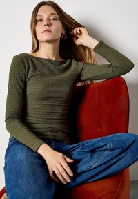 Olive green long-sleeve top with textured fabric, fitted design, and round neckline, paired with blue jeans, seated on a red velvet chair.