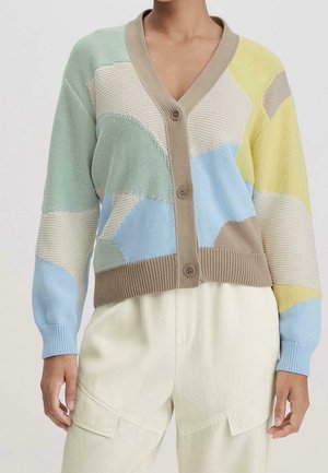 Personne portant un cardigan tricoté patchwork multicolore avec des boutons, associé à un pantalon couleur crème, visible du cou aux hanches.