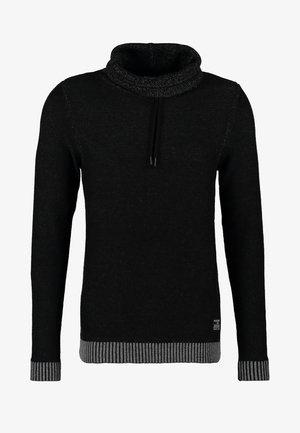 Uomo che indossa un maglione nero e jeans grigi, tiene in mano una borsa in pelle grigia, in piedi davanti a uno sfondo semplice.