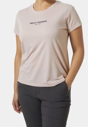 Personne portant un t-shirt rose clair Helly Hansen et un pantalon gris foncé avec une main dans la poche, sur un fond uni.