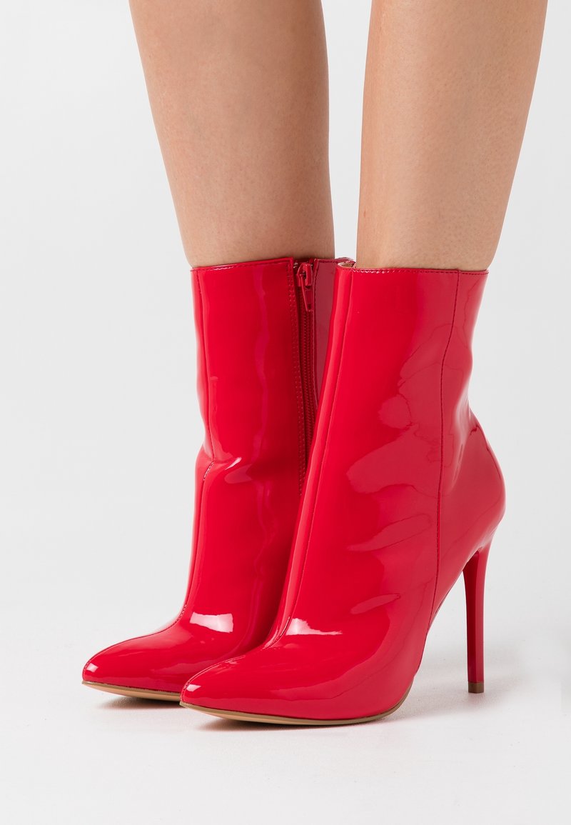 Bottines vernies rouges à talons hauts avec bouts pointus et fermetures éclair latérales portées sur jambes nues devant un fond blanc.