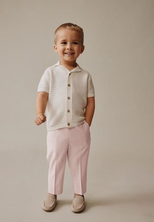 Junger Junge steht und lächelt, trägt ein weißes kurzärmeliges Hemd mit Knöpfen, hellrosa Hose und beige Schlupfschuhe.