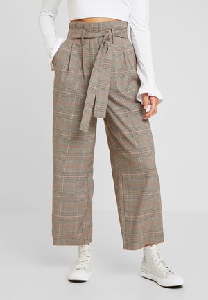 Kvinde iført viddebenede ternede bukser med højtaljetalje og bindebælte, hvid crop top med lange ærmer og hvide high-top sneakers.