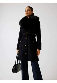 Abrigo negro con cuello de pelo sintético, botones dorados y un cinturón. Cuenta con dos bolsillos delanteros y se combina con pantalones vaqueros oscuros y un bolso negro.