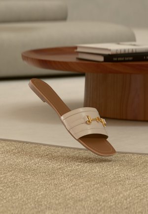 Beżowy skórzany sandał wsuwany z detalem w postaci złotej klamry, spoczywający na beżowym, teksturowanym dywanie obok drewnianego, okrągłego stolika kawowego z poukładanymi książkami.