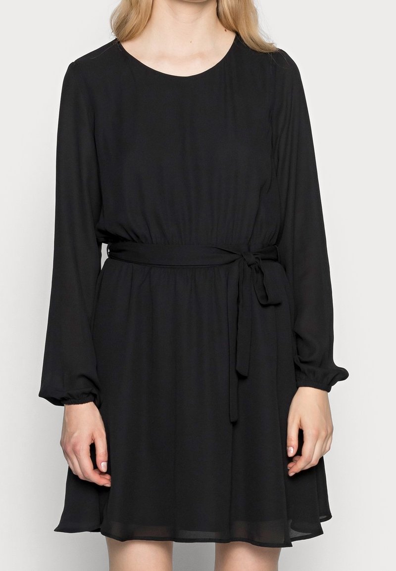 Robe noire à manches longues en tissu léger, présentant une taille cintrée avec un lien et une jupe fluide. Texture lisse et col rond.