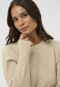 Joven mujer con cabello largo y oscuro, vestida con un suéter de punto beige, tocándose suavemente la barbilla con la mano izquierda frente a un fondo liso.