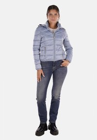 Giacca trapuntata blu chiaro con cappuccio, chiusura lampo frontale e due tasche laterali. Indossata con jeans blu scuro e stivali neri robusti.