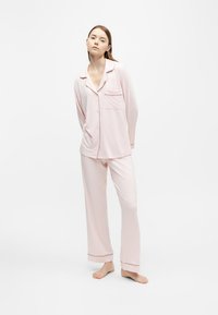 Femme debout pieds nus portant un haut de pyjama rose clair à manches longues avec boutons et un pantalon de pyjama large assorti sur fond blanc.
