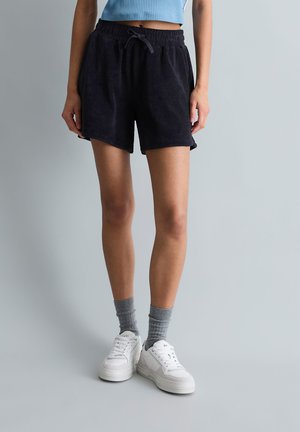 Vrouw draagt zwarte shorts met trekkoord, grijze sokken, witte sneakers en een lichtblauw cropped topje tegen een effen grijze achtergrond.
