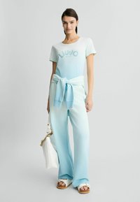 T-shirt bleu clair avec "LIU JO MILANO" imprimé dans une teinte plus foncée, associé à un pantalon large bleu pâle assorti et des sandales blanches.