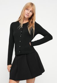 Cárdigan negro de manga larga hecho de tela suave, con escote redondo, cierre frontal de botones y un diseño ajustado combinado con una falda negra plisada.