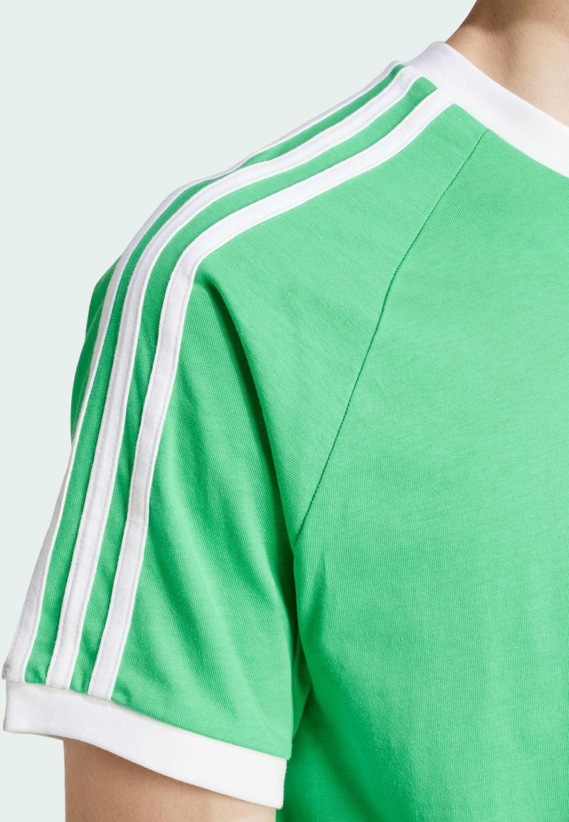 Camiseta de algodón verde con cuello redondo de canalé blanco y tres rayas blancas que recorren los hombros. Textura suave, ajuste casual.