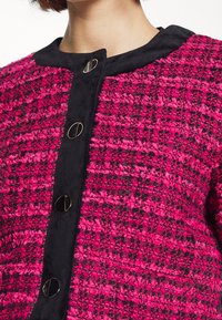 Vävd rosa och svart jacka med en texturerad yta, rund halsringning och svarta knapplås framtill.