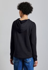 Schwarzer Kapuzenpullover mit langen Ärmeln, aus weichem Stoff hergestellt. Merkmale ein einfaches Design, eine lässige Passform und gerippte Bündchen, kombiniert mit blauen Jeans.