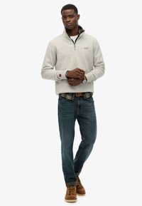 Jasnoszara bluza na zamek, dżinsowe spodnie i brązowe buty. Założona na koszulę w kratę. Z logo na piersi i z podwiniętymi rękawami.