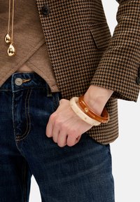 Drei Armreifen aus unterschiedlichen Materialien: einer aus Bernstein, einer weiß mit einem Sonnendesign und einer braun, getragen an einem Handgelenk gegen Denim.