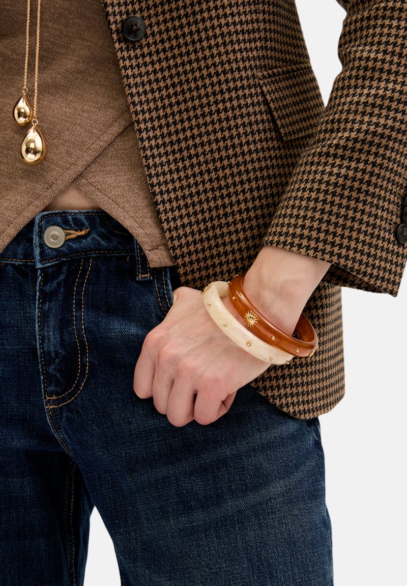 Drei Armreifen aus unterschiedlichen Materialien: einer aus Bernstein, einer weiß mit einem Sonnendesign und einer braun, getragen an einem Handgelenk gegen Denim.