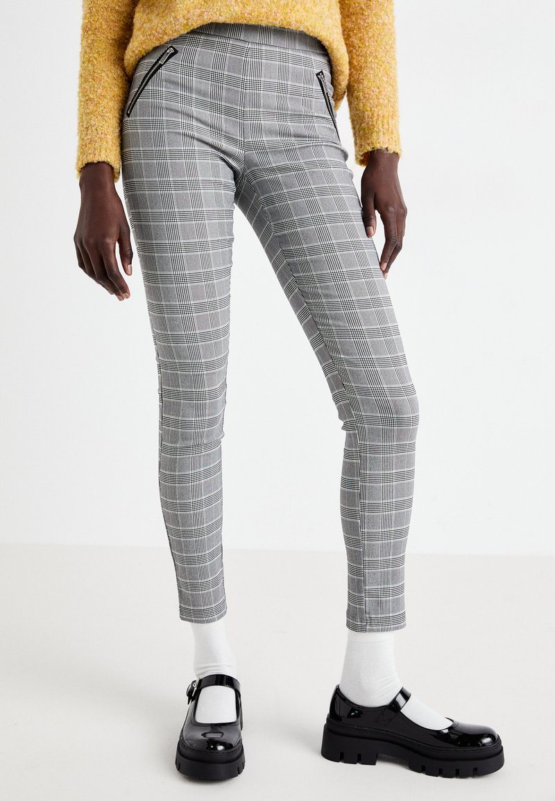 Pantalon à carreaux gris avec une coupe slim, doté de zips sur les côtés, associé à des chaussures noires et des chaussettes blanches. Le tissu a une texture lisse.