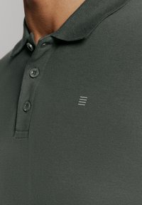 Donkergroene poloshirt met gestructureerde stof, twee knoppen bij de kraag en een subtiele logo-detail met drie lijnen op de borst.