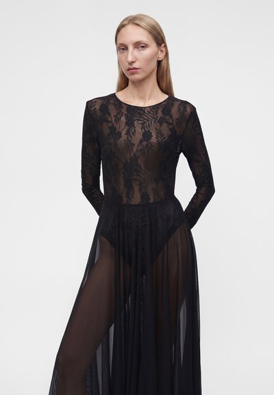 Ξανθιά γυναίκα με μαύρο, μακρυμάνικο, διάφανο δαντελένιο φόρεμα με λουλούδια, που στέκεται μπροστά σε απλό ανοιχτό φόντο.