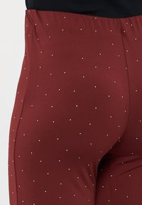 Burgundy leggings dengan tekstur halus, dihiasi dengan studs dekoratif kecil berwarna putih yang tersebar merata di seluruh kain. Desain pinggang tinggi.