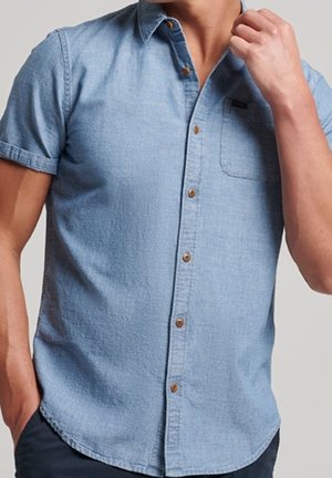 Mand iført lyseblå kortærmet skjorte med brune knapper, den ene hånd justerer kraven, den anden hånd i lommen.