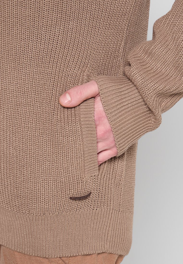 Beige gestrickter Pullover mit einem strukturierten Muster, ausgestattet mit gerippten Bündchen und einer Seitentasche. Kleines Logo-Detail in der Nähe des Saums.