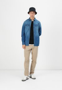 Jeans košulja u plavoj boji s džepovima na prsima, složena preko crne majice. Bež hlače i crni šešir sa širokim obodom upotpunjuju izgled. Ležerna obuća.